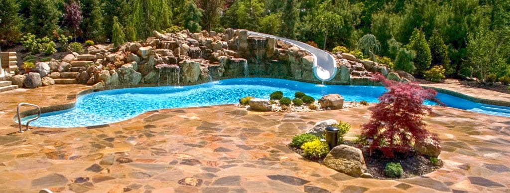 Rock waterfall with integrated water slide on a large backyard swimming pool.