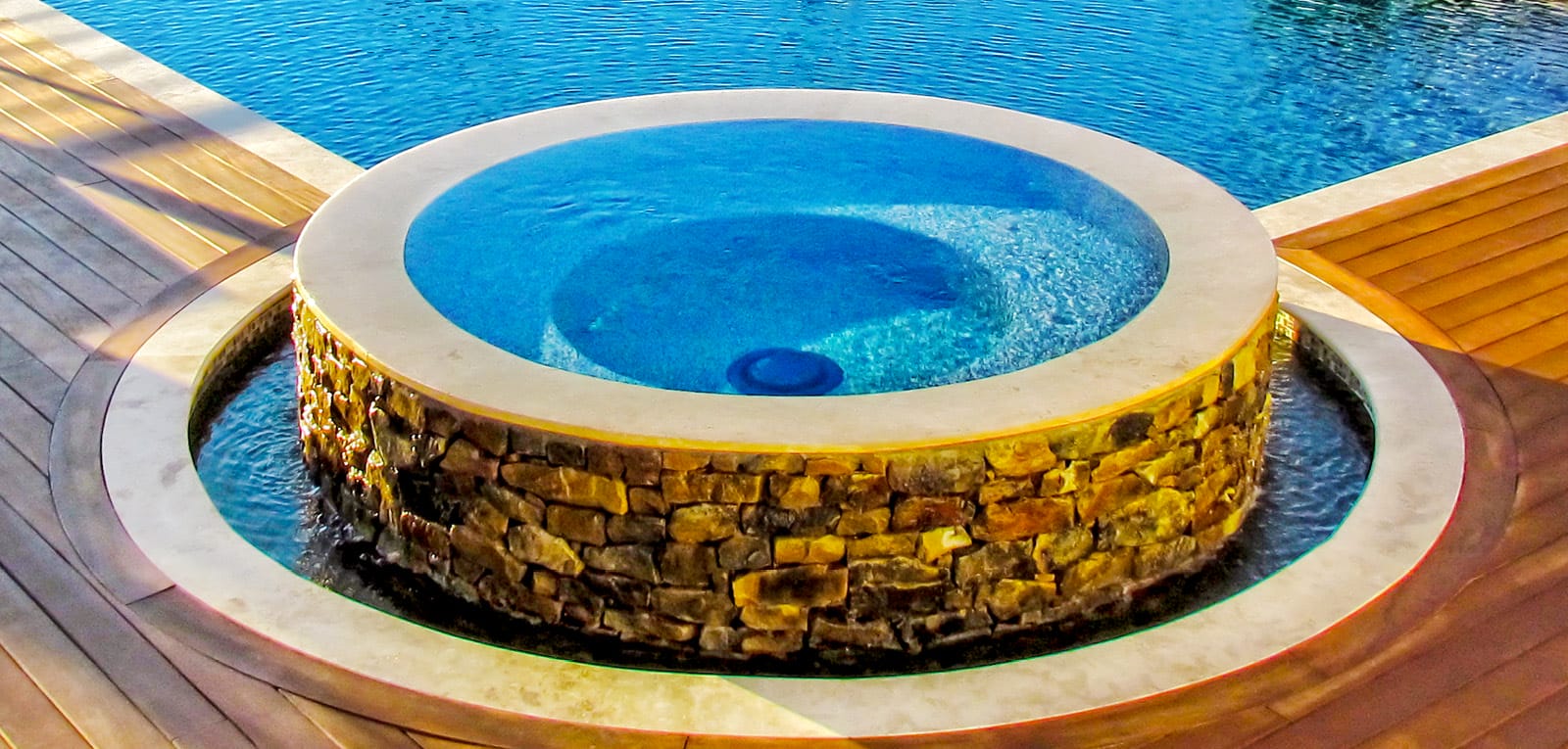 Round rimflow spa with rustic stone exterior on inground pool.