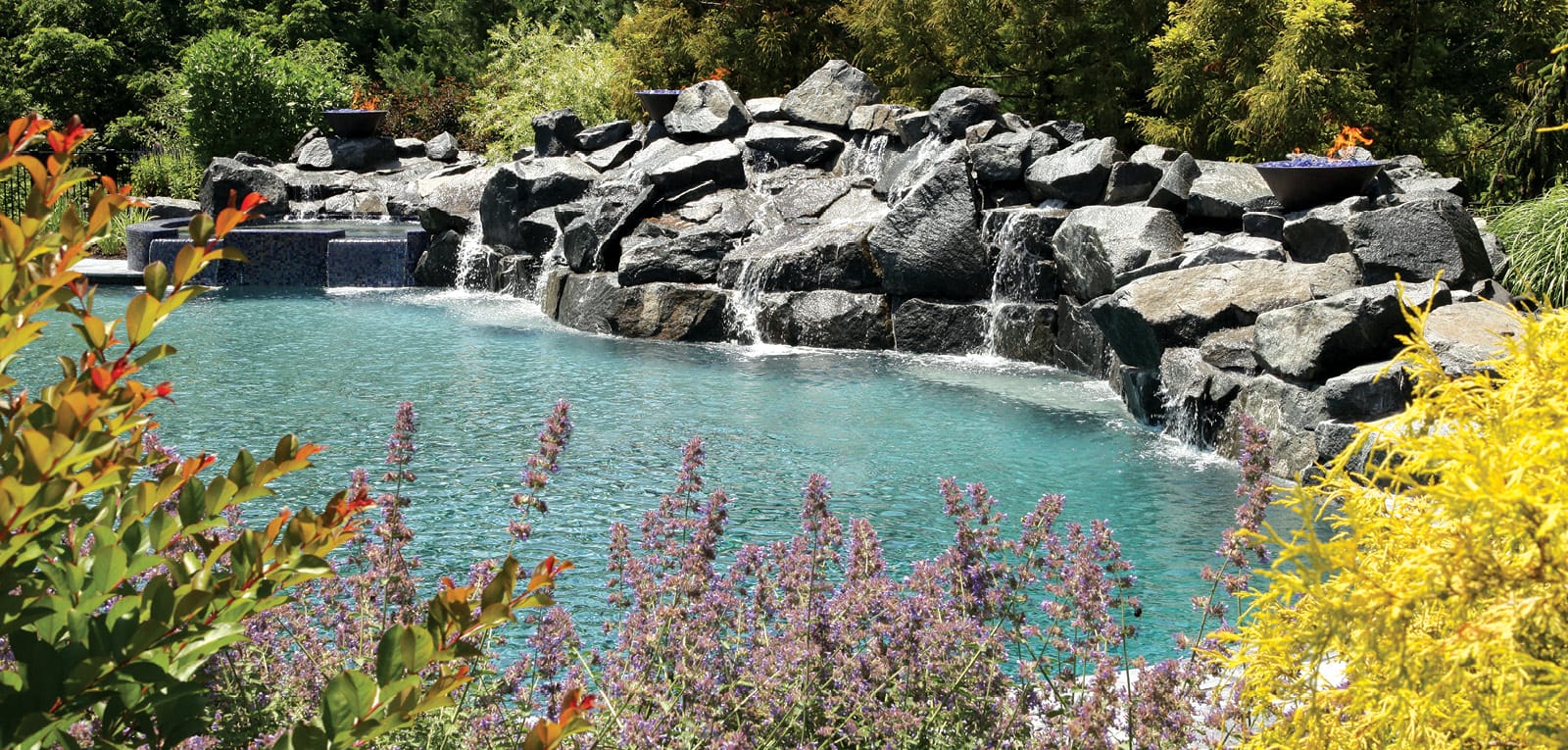 Large, sprawling stone waterfall flanked with fire bowls on a naturalistic swimming pool.