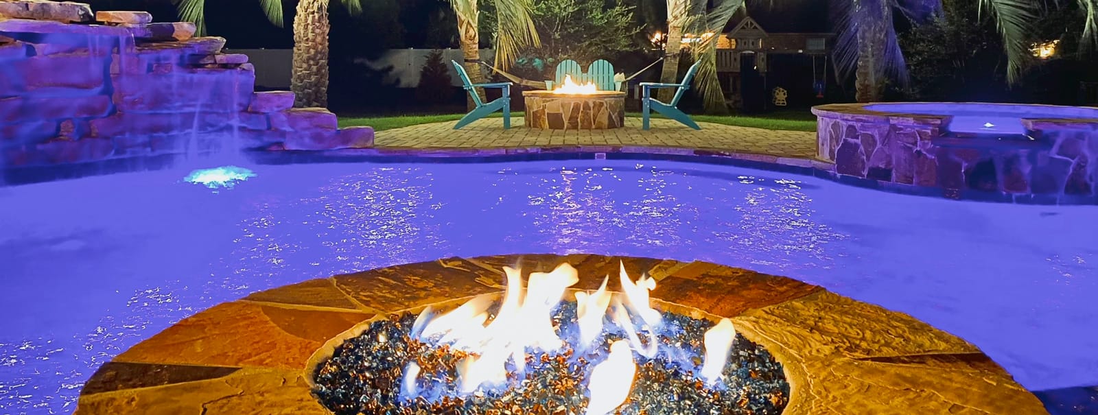 Fire pit in front of Jacksonville gunite pool with purple illumination at night.