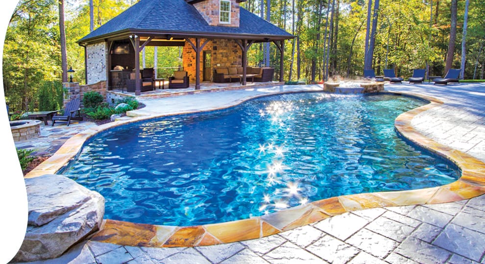 Curvy swimming pool with water sparkling in the sunlight in a wooded backyard..