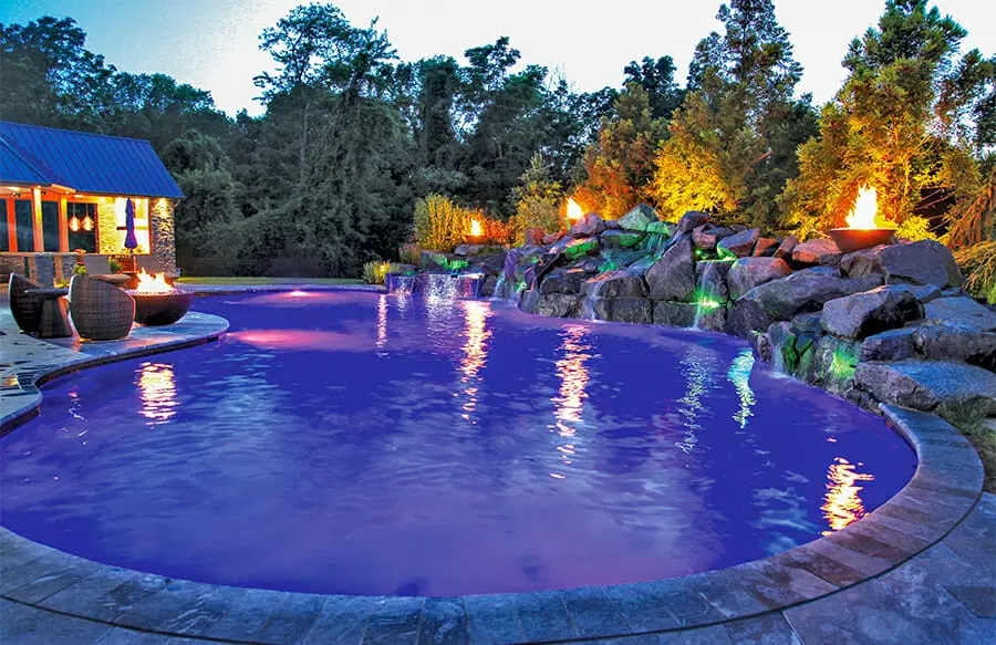 Free-form pool at night with stone waterfall and fire bowls.