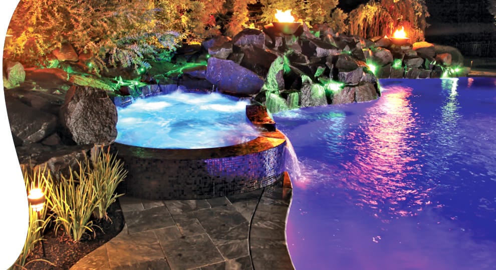 Swimming pool and spa with rock waterfall and fire features illuminated in purple and green light at night.