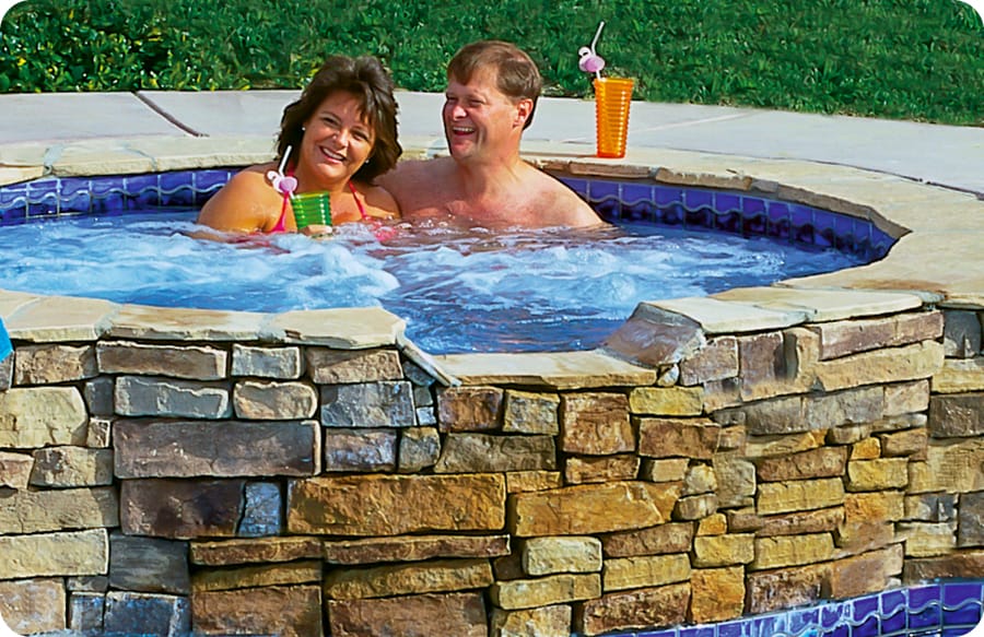 Man and woman couple with drinks enjoying bubbling water in a stone-faced swimming pool spa.