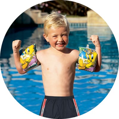 Young buy wearing flotation devices on his arms smiling in front of swimming pool. 