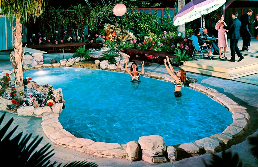 Mid-Century era swimmers playing with inflatable ball at night in lagoon-style swimming pool with rock perimeter.