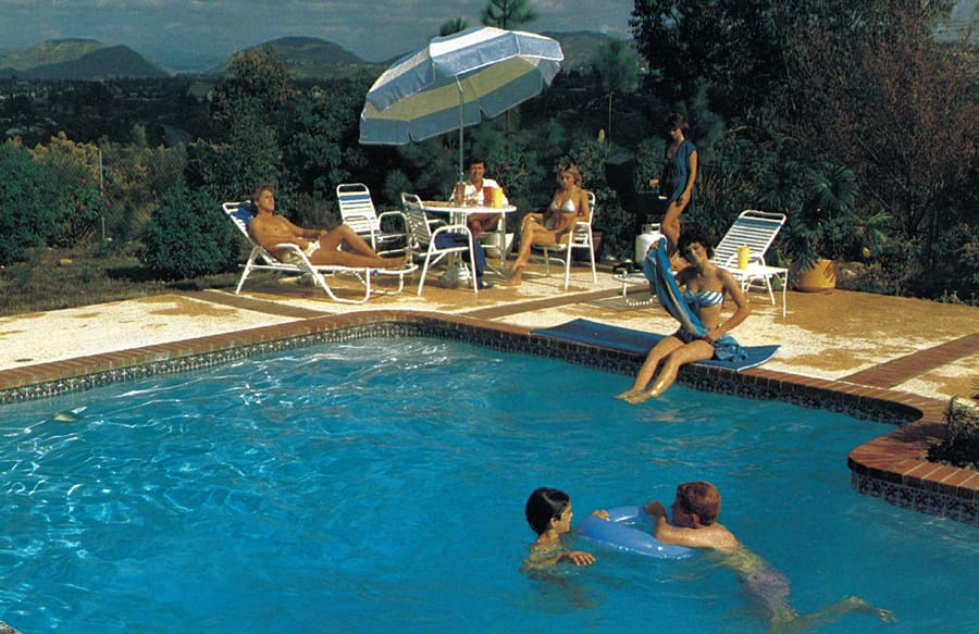 Kids playing in an early 1980s swimming pool with adults sitting at tables on the deck.
