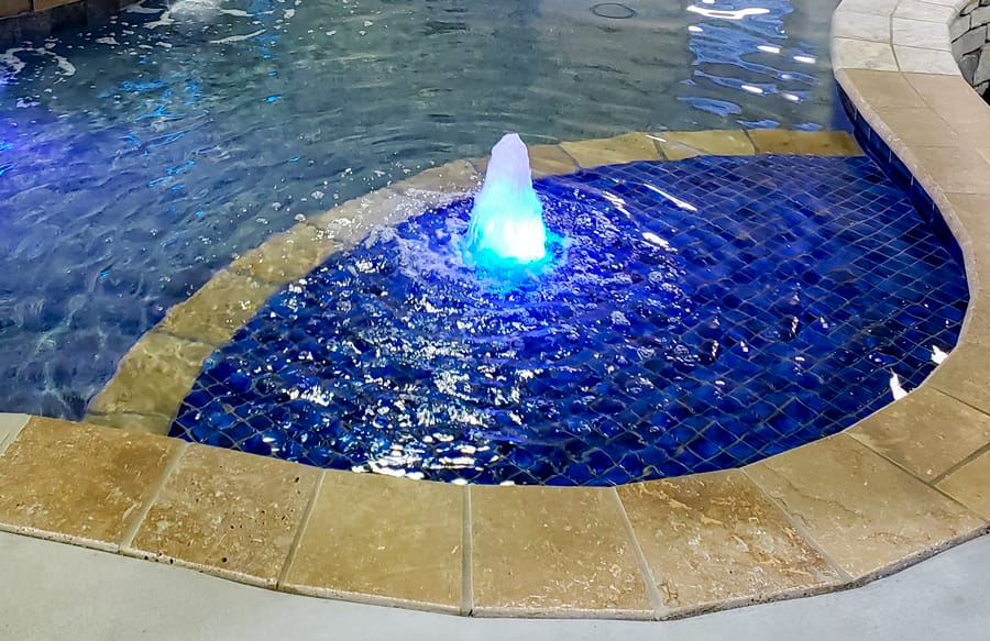 Swimming pool tanning ledge covered in blue tile with illuminated fountain.