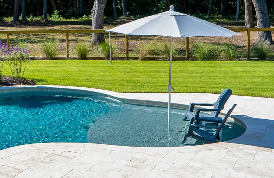 Tanning ledge with two upright chairs and white patio umbrella on swimming pool.