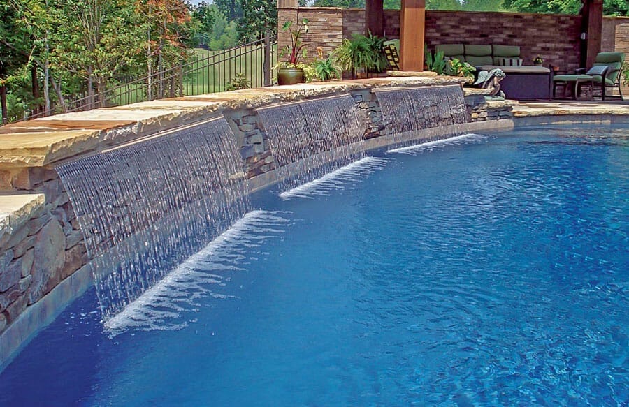 Trio of teardrop-style cascade water features on raised wall of swimming pool.