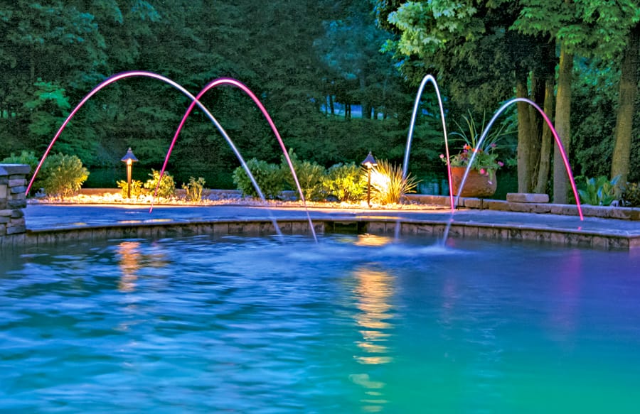 Four illuminated arcing water laminars over inground pool.