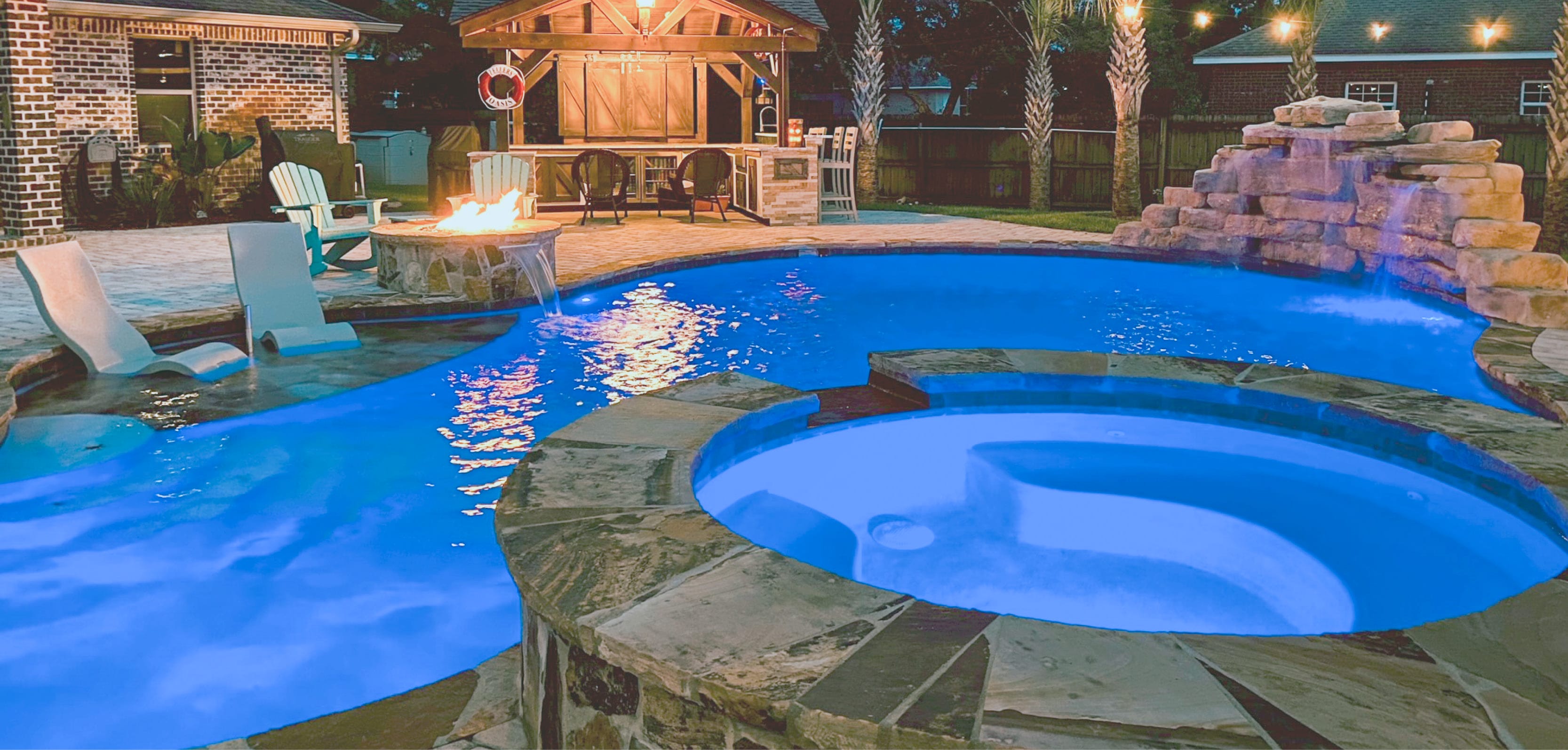 Residential pool with a spa, waterfall, and fire pit, illuminated in blue light in a backyard.