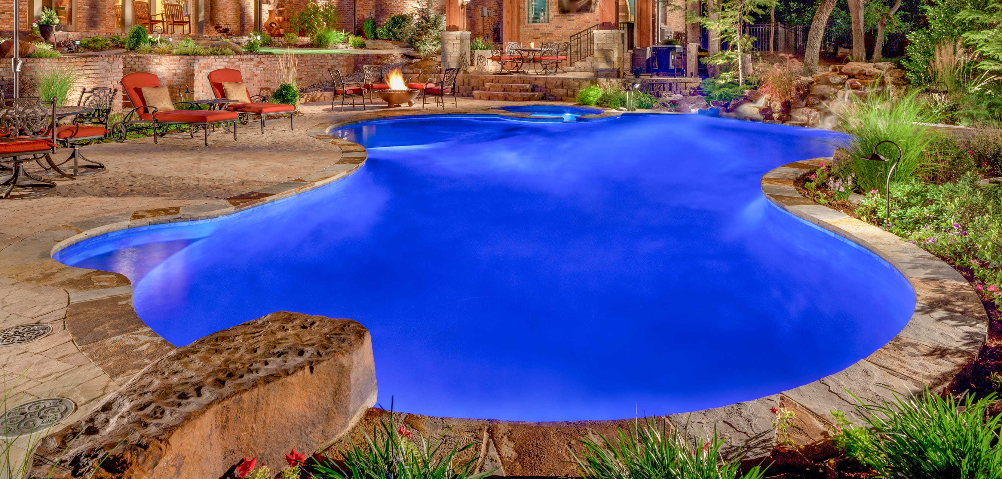Curvy gunite pool illuminated at night in a woodsy backyard.